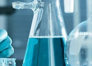 A close-up of a laboratory flask filled with blue liquid, alongside another glass container. A gloved hand is visible on the left, suggesting a scientific or laboratory setting.