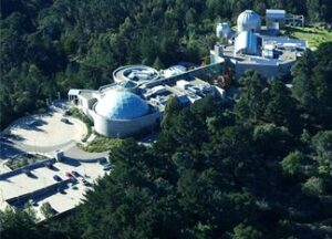 Aerial view of a science museum complex with domed and modern buildings surrounded by dense green trees, featuring parking areas and winding roads.
