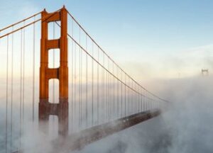 The Golden Gate Bridge towers rise above dense fog, with the bridge partially obscured and the sky clear above.