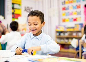 A young boy smiles while playing with building blocks at a table in a colorful classroom. Other children and educational materials are visible in the background.