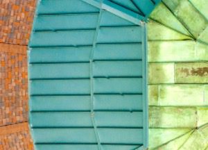 Close-up, top-down view of a domed roof showing three distinct sections: red-brown shingles, blue-green metal panels, and weathered green copper panels, divided by lines and different textures.