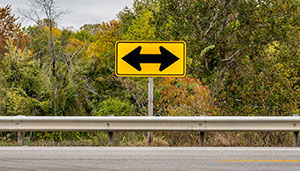 A yellow road sign with black arrows redirects drivers left and right, standing by a roadside with a metal guardrail and trees in the background.