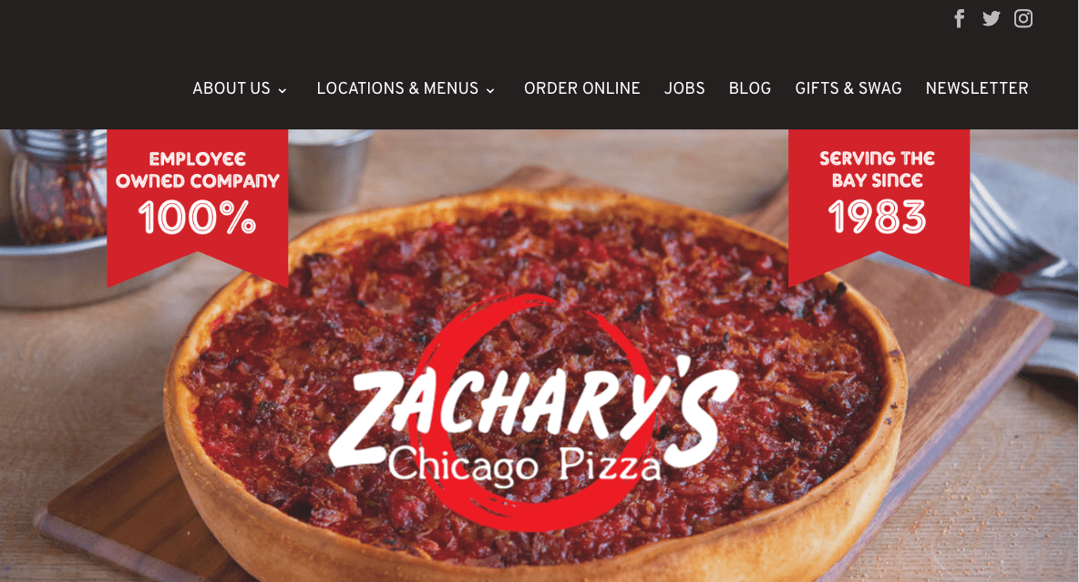 A deep dish pizza topped with tomato sauce sits on a wooden table. Banners above read Employee Owned Company 100% and Serving the Bay Since 1983. The Zachary’s Chicago Pizza logo is centered over the pizza.