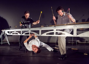 Three men play an unusual metal percussion instrument. Two stand above, striking it with mallets, while one crouches below, also playing, with dramatic lighting highlighting their performance.