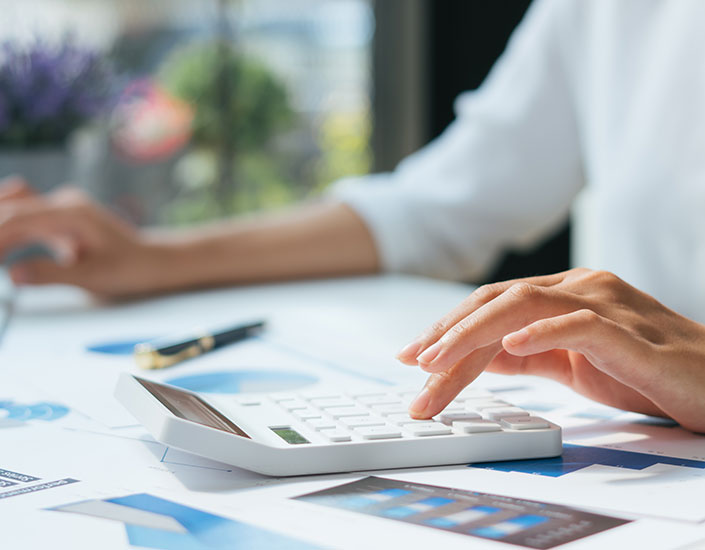 Close-up of a person’s hand pressing buttons on a white calculator, with documents, charts, and a pen on the desk, suggesting financial calculations or office work.