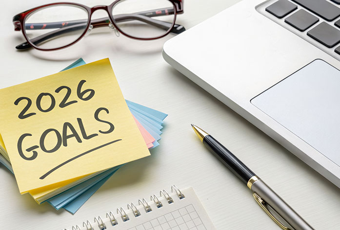 A sticky note reading 2026 GOALS sits on a desk next to a laptop, pen, spiral notebook, and eyeglasses, suggesting planning and goal setting.