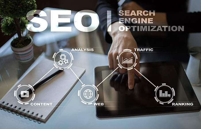 A person points to a tablet screen on a desk with a plant, notebook, and pen. Overlay text reads “SEO Search Engine Optimization” and highlights concepts: content, analysis, web, traffic, and ranking with icons.
