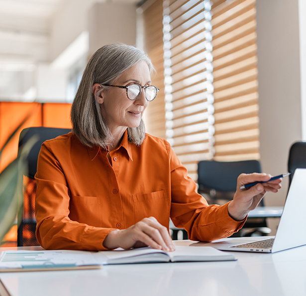 An older woman with gray hair and glasses, wearing an orange shirt, sits at a desk in a bright office, smiling and pointing at her laptop screen while taking notes in a notebook.