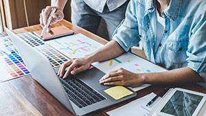 Two people work at a desk with a laptop, color swatches, printed charts, and a tablet. One person types while the other points to a document, suggesting a collaborative design or planning process.