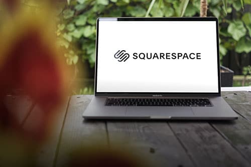 An open laptop displaying the Squarespace logo sits on an outdoor wooden table with green plants in the blurred background.